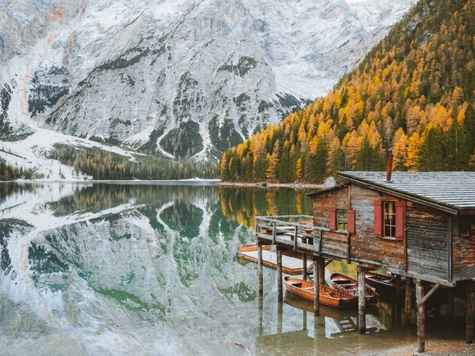 בקתת עץ על אגם עם השתקפות הרים מושלגים ועצי שלכת בדולומיטים