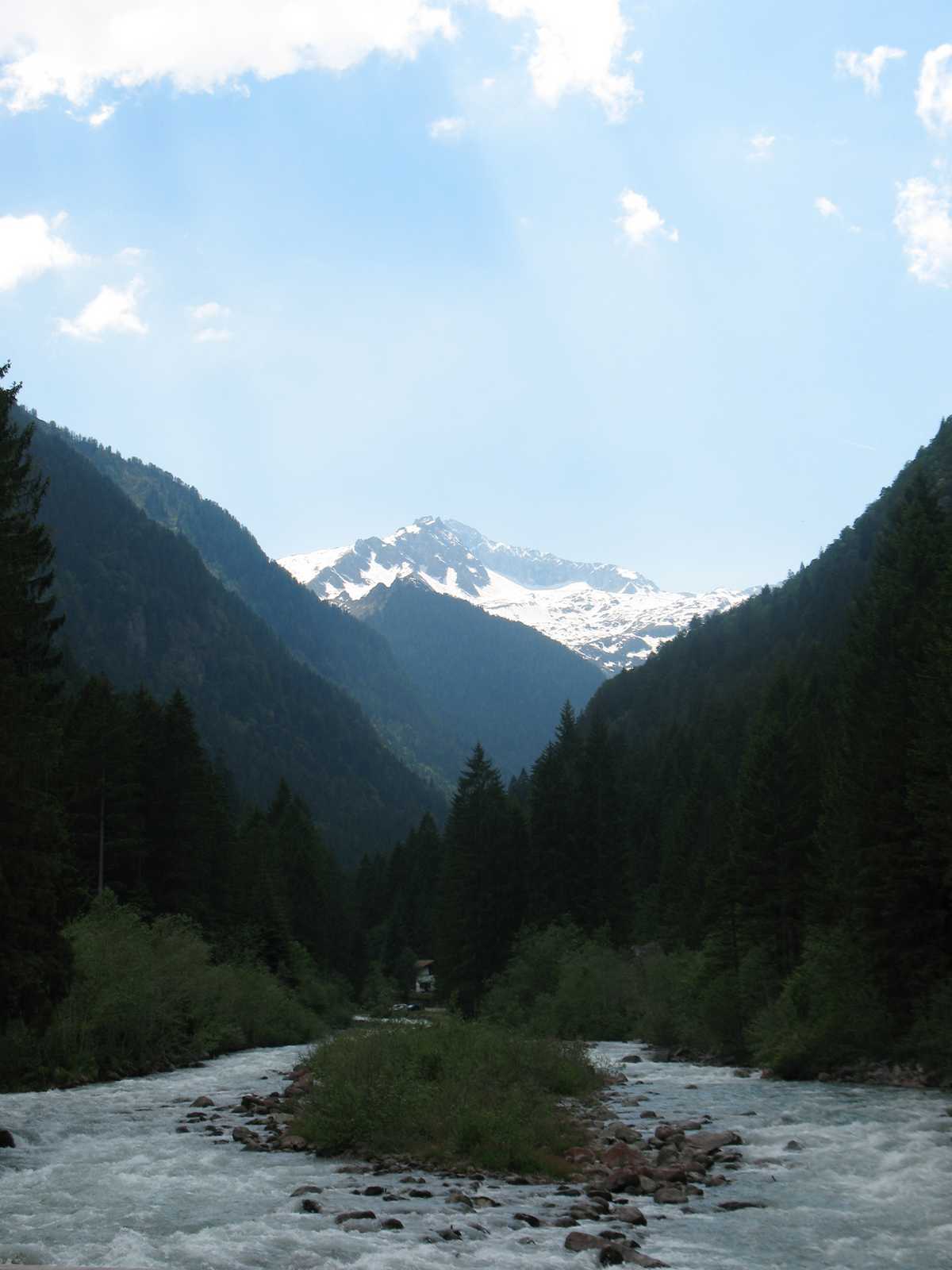 עמק הררי עמוק באזור דולומיטים עם נהר זורם ופסגה מושלגת באופק.