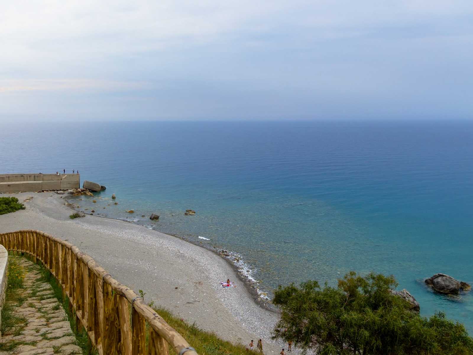 מבט מלמעלה על חוף ים בקלבריה עם גדר עץ ומים בצבע טורקיז תחת שמיים מעוננים.