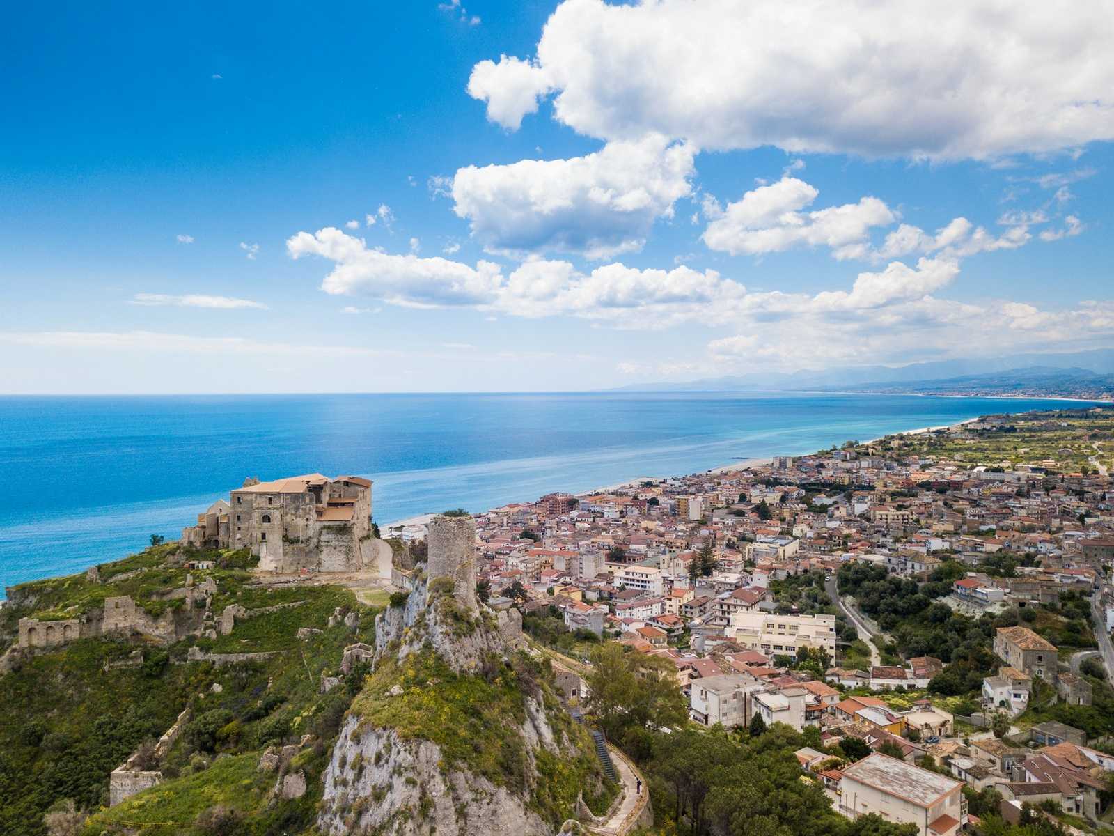 נוף פנורמי של עיר חוף וטירה עתיקה בקלבריה עם גגות רעפים אדומים.